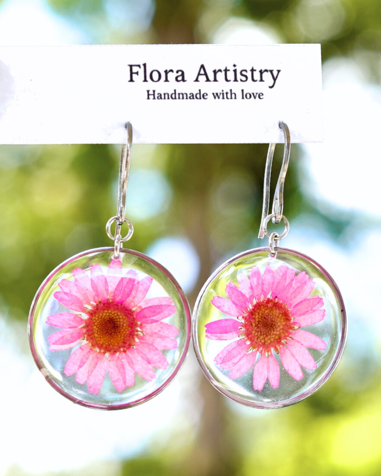 Floral earrings with pink flowers in a clear setting against a blurred natural background.